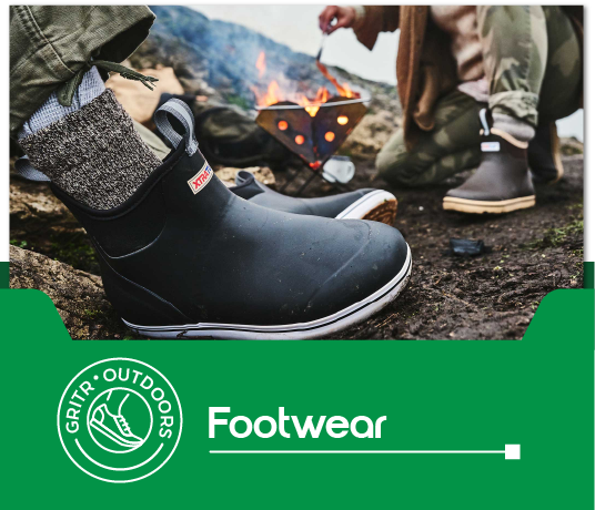 graphic, An individual wearing black waterproof boots with a knitted grey cuff, standing on rocky terrain next to a small, portable campfire. Another person is visible in the background tending to the fire. text, footwear